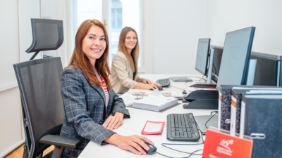 Zwei Frauen sitzen an Schreibtischen mit Computern und arbeiten in einem modernen Büroumfeld.