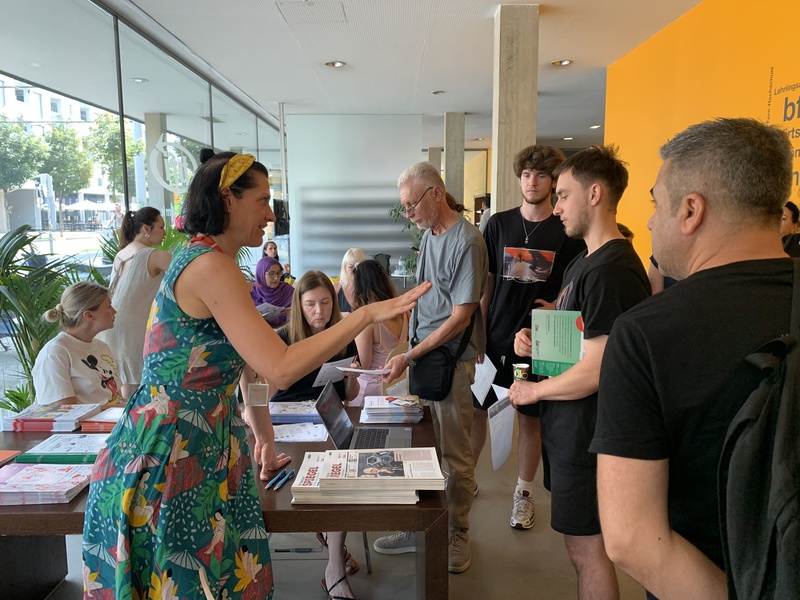 Eine Frau im bunten Kleid spricht mit einer Gruppe von Menschen an einem Informationsstand.