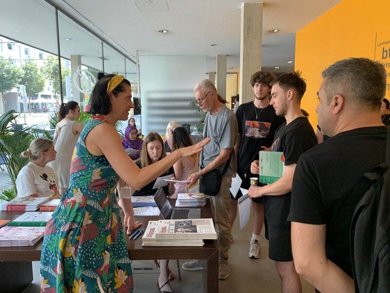 Eine Frau im bunten Kleid spricht mit einer Gruppe von Menschen an einem Informationsstand.