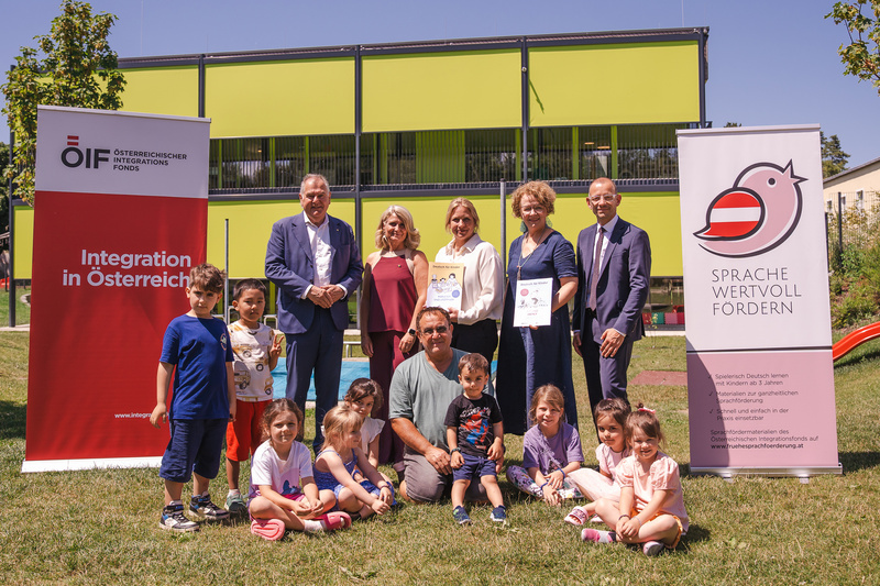Gruppenfoto von Erwachsenen und Kindern vor zwei Werbebannern für Integration und Sprachförderung in Österreich.