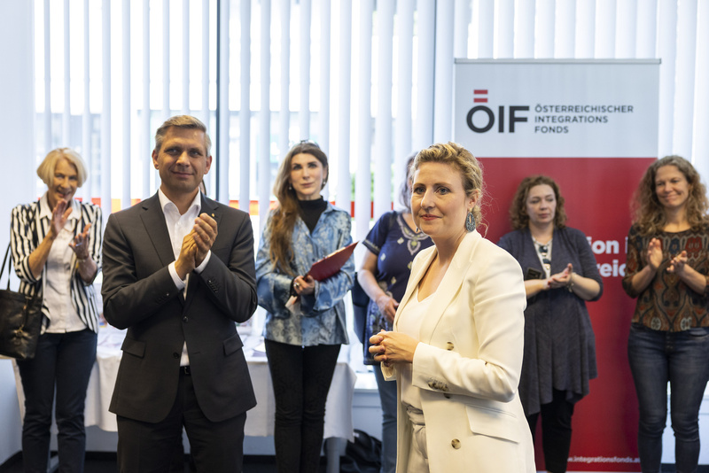 Die Veranstaltung zeigt applaudierende Menschen, im Vordergrund eine Frau in weißem Anzug, die angesprochen wird.