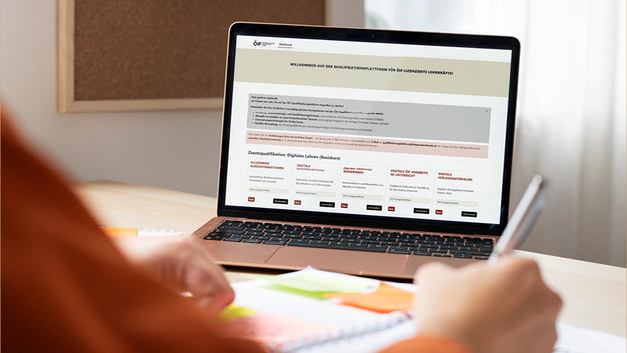 Eine Person sitzt an einem Tisch und nutzt einen Laptop. Der Bildschirm zeigt eine Webseite mit Informationen zu einem Programm oder einer Dienstleistung. Auf dem Tisch liegen Notizblätter und ein Stift, während ein Korkbrett im Hintergrund sichtbar ist.