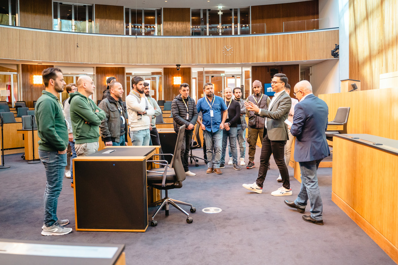 Eine Gruppe von Menschen hört einem Redner in einem Raum mit Tischen und Stühlen zu.