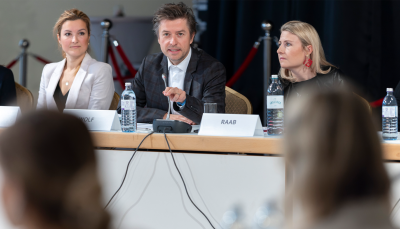 Drei Personen sitzen an einem Tisch während einer Diskussion oder Pressekonferenz.