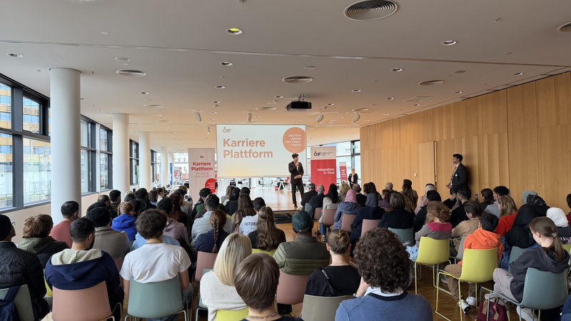 Eine Gruppe von Menschen sitzt auf Stühlen in einem hellen Raum. Auf einer Bühne spricht ein Referent vor einem großen Bildschirm mit der Aufschrift „Karriere Plattform“. Die Zuschauer scheinen aufmerksam zuzuhören.