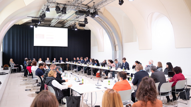 Eine große Gruppe von Menschen sitzt um einen Tisch in einem Konferenzraum und diskutiert.