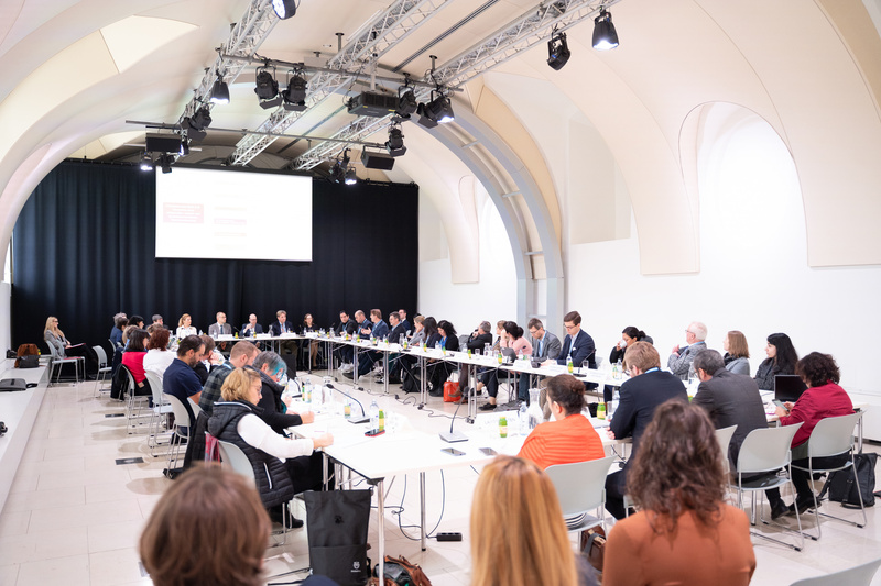 Eine große Gruppe von Menschen sitzt um einen Tisch in einem Konferenzraum und diskutiert.