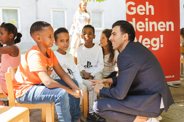 Ein Mann im Anzug spricht mit mehreren Kindern in einem sonnigen, offenen Bereich.