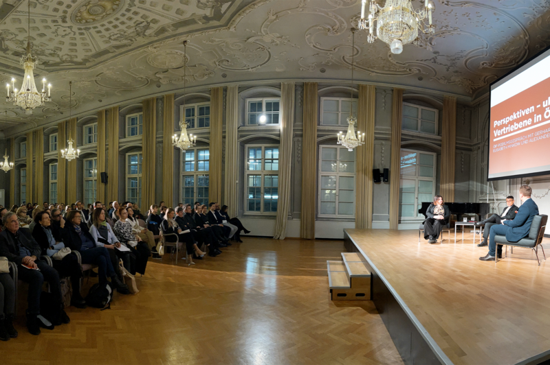 Podiumsdiskussion mit Teilnehmern zu Themen der Perspektiven und Vertreibung in Österreich. Publikum sitzt aufmerksam im Saal.
