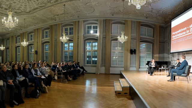 Podiumsdiskussion mit Teilnehmern zu Themen der Perspektiven und Vertreibung in Österreich. Publikum sitzt aufmerksam im Saal.