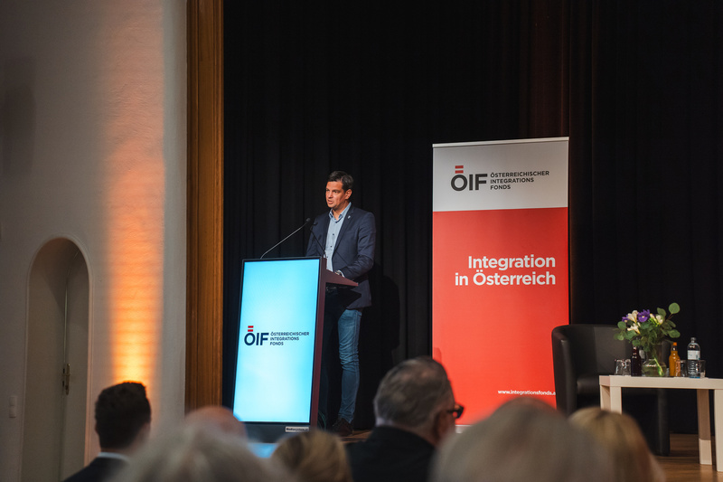 Ein Mann spricht an einem Rednerpult vor einem Banner über Integration in Österreich.