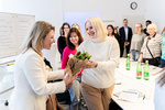 Eine Frau überreicht einer anderen Frau einen Blumenstrauß in einem Raum mit mehreren Personen.
