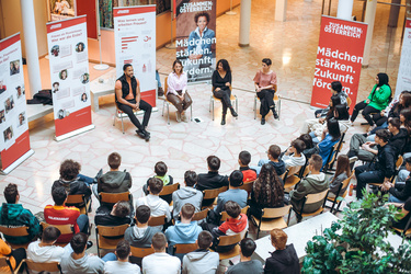 Podiumsdiskussion mit vier Sprechern, Zuschauer in einem modernen Raum, Fokus auf Frauenförderung und Bildung.