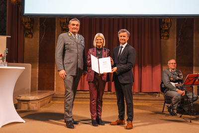 Drei Personen stehen auf einer Bühne und halten eine Urkunde während einer Zeremonie.