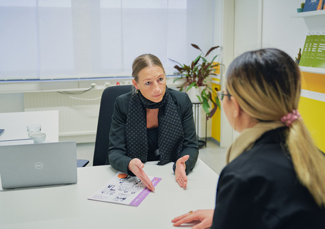 Zwei Frauen führen ein Gespräch in einem modernen Büro, eine erklärt etwas mit ausladenden Gesten.
