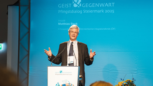 Ein Mann spricht auf einer Bühne bei einer Veranstaltung mit blauem Hintergrund.