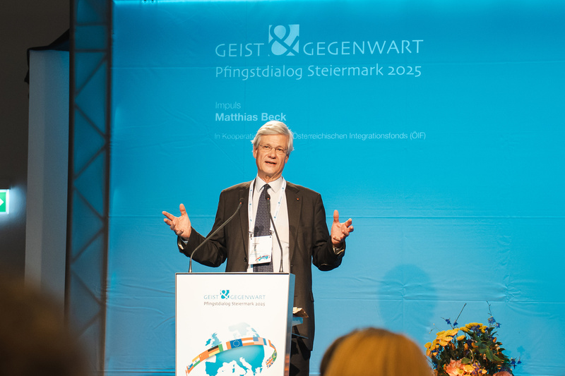 Ein Mann spricht auf einer Bühne bei einer Veranstaltung mit blauem Hintergrund.