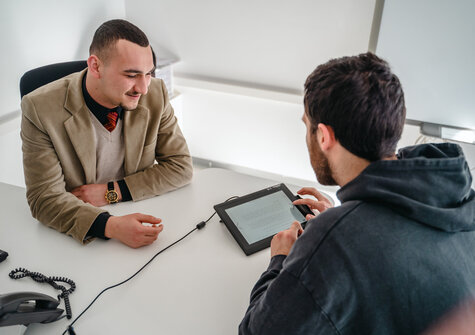 Zwei Männer sitzen an einem Tisch und diskutieren, während einer auf ein Tablet blickt.