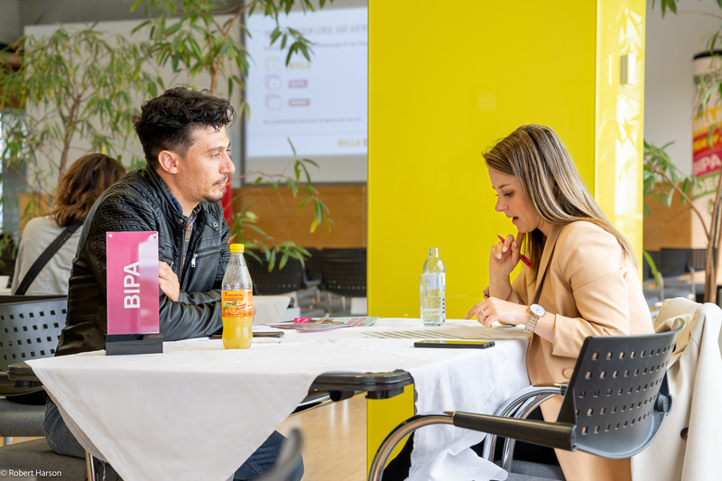 Zwei Personen sitzen an einem Tisch, diskutieren und arbeiten an Dokumenten. Ein Schild mit "BIPA" ist sichtbar.