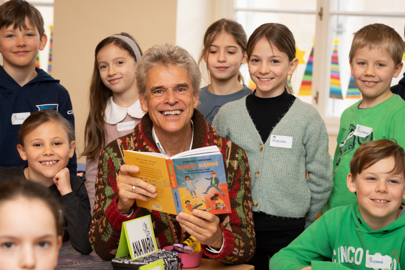 Ein lächelnder Mann liest Kindern ein Buch vor, umgeben von mehreren aufmerksamen, fröhlichen Kindern.