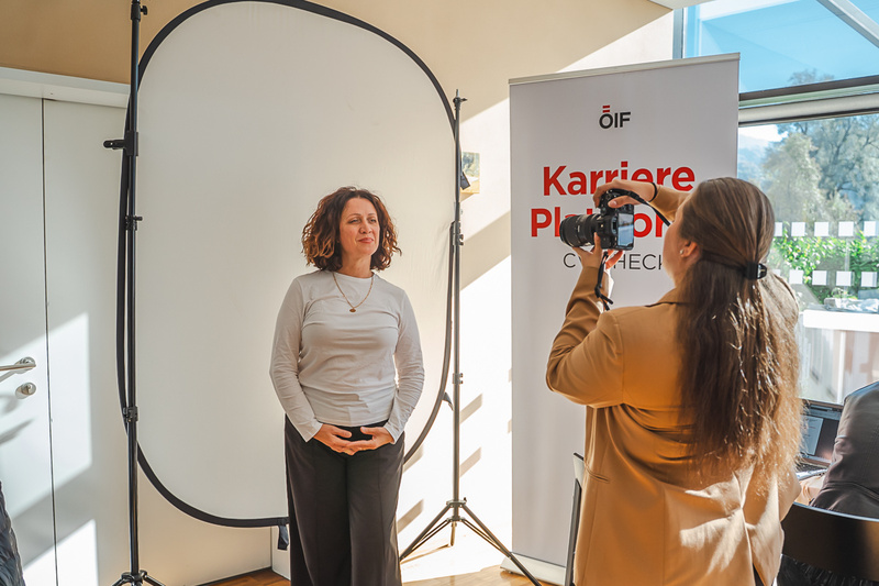 Eine Frau posiert für ein Foto vor einer neutralen Kulisse, während eine andere Person mit einer Kamera fotografiert.