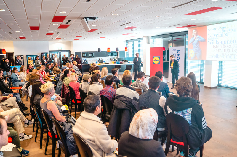 Eine Präsentation vor Publikum mit zwei Rednern, das Begrüßungen in verschiedenen Sprachen zeigt.