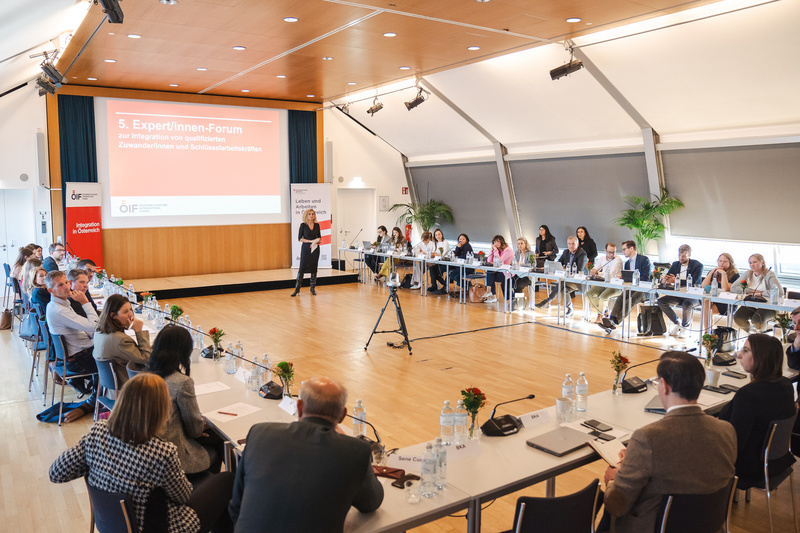 Ein Expert:innen-Forum zur Integration qualifizierter Zuwanderer mit Vortrag und Teilnehmern an Tischreihen.