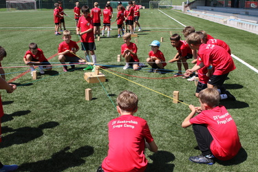 Kinder in roten Trikots arbeiten zusammen, um ein Teamspiel mit Seilen und Holzblöcken auf einem Sportplatz zu lösen.
