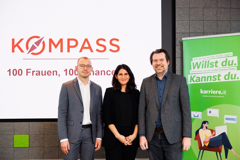 Drei Personen stehen vor einem Banner mit dem Schriftzug "KOMPASS: 100 Frauen, 100 Chancen".