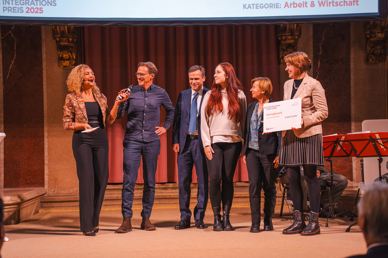 Eine Frau spricht auf der Bühne, während fünf Personen eine Auszeichnung für den Integrationspreis 2025 erhalten.