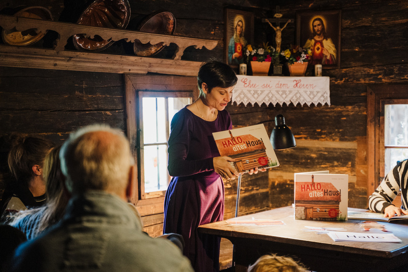Eine Frau präsentiert ein Buch mit dem Titel „Hallo, altes Haus!“ vor einer Gruppe von Zuhörern.
