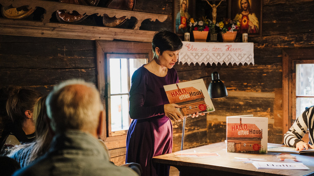Eine Frau präsentiert ein Buch mit dem Titel „Hallo, altes Haus!“ vor einer Gruppe von Zuhörern.