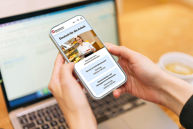 Eine Person hält ein Smartphone, auf dem eine Webseite mit dem Titel „Deutsch für die Arbeit“ angezeigt wird. Im Hintergrund ist ein Laptop sichtbar. Die Webseite zeigt Informationen zu einem Serviceangebot und den Öffnungszeiten.