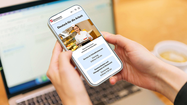 Eine Person hält ein Smartphone, auf dem eine Webseite mit dem Titel „Deutsch für die Arbeit“ angezeigt wird. Im Hintergrund ist ein Laptop sichtbar. Die Webseite zeigt Informationen zu einem Serviceangebot und den Öffnungszeiten.