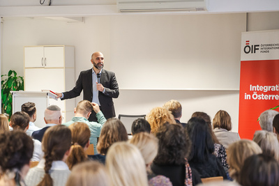 Ein Mann spricht vor einem Publikum in einem Raum, im Hintergrund steht ein Plakat zur Integration in Österreich.