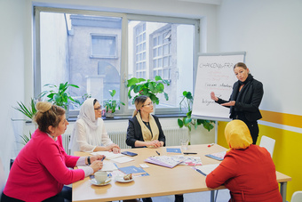 Eine Frau erklärt während eines Meetings in einem hellen Raum an einem Flipchart, umgeben von vier weiteren Frauen.