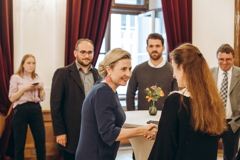 Zwei Frauen schütteln sich die Hände, während andere Personen lächeln und zuschauen. Ein Blumenarrangement ist sichtbar.