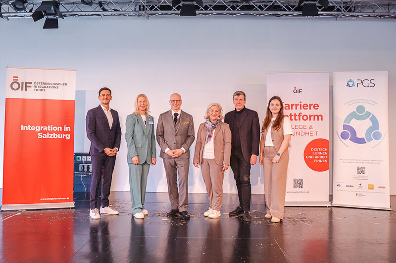 Eine Gruppe von sechs Personen steht auf einer Bühne, umgeben von Banner mit Aufschriften zur Integration und Karriereplattform in der Pflege und Gesundheit. Sie wirken förmlich gekleidet und präsentieren sich gemeinsam für ein Foto.