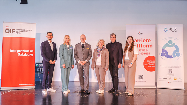 Eine Gruppe von sechs Personen steht auf einer Bühne, umgeben von Banner mit Aufschriften zur Integration und Karriereplattform in der Pflege und Gesundheit. Sie wirken förmlich gekleidet und präsentieren sich gemeinsam für ein Foto.