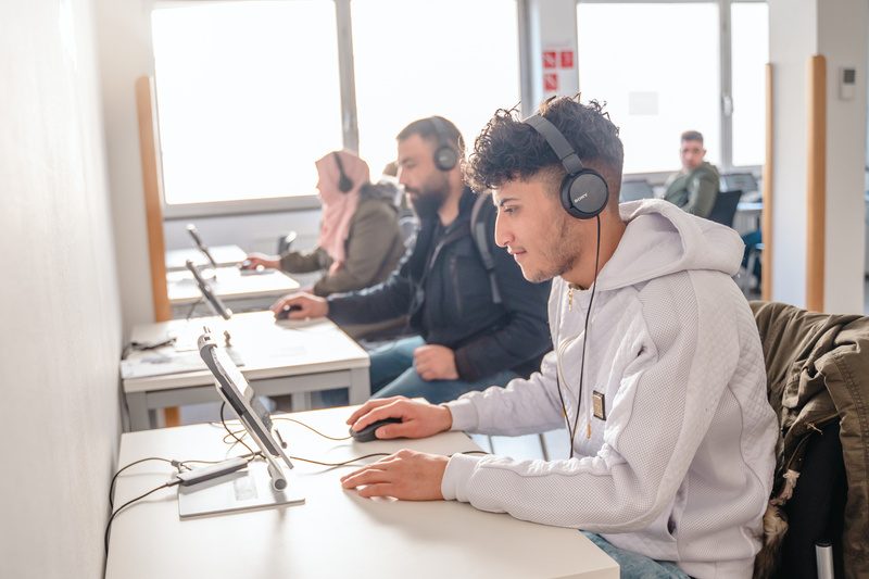 Studenten sitzen an Tischen mit Headsets und arbeiten konzentriert an Tablets in einem hellen Raum.