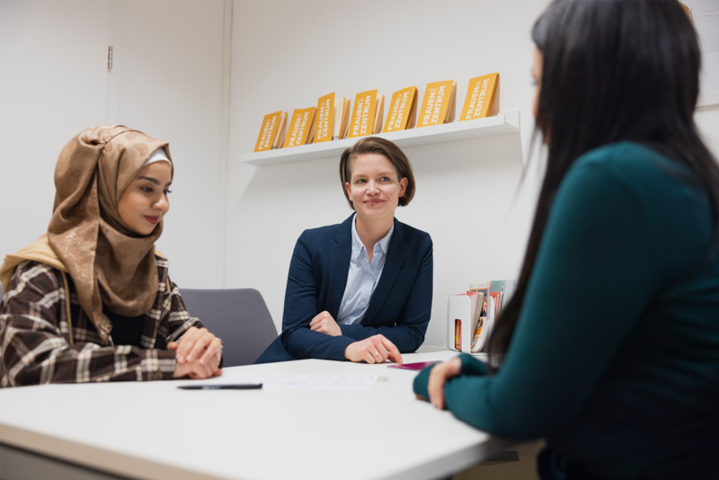 Drei Frauen sitzen an einem Tisch in einem Büro und führen ein Gespräch.