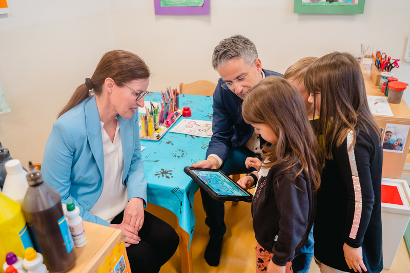 Eine Frau und ein Mann zeigen Kindern auf einem Tablet etwas. Der Raum ist kreativ und kinderfreundlich gestaltet.