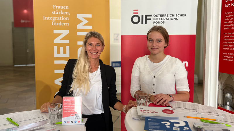 Zwei Frauen an einem Informationsstand, der Integration und Frauenförderung thematisiert.