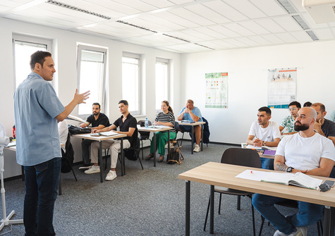 Ein Lehrer steht vor einer Gruppe von Schülern in einem Klassenzimmer. Alle hören aufmerksam zu.