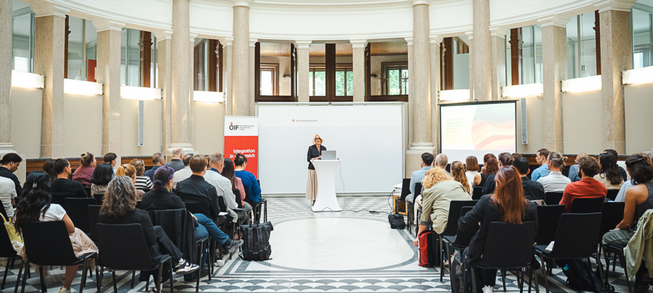 Eine Person steht auf einer Bühne in einem saal mit hohen Decken, umgeben von Säulen. Vor ihr sitzt ein Publikum, das auf die Präsentation schaut. Der Raum hat große Fenster und eine elegante, symmetrische Gestaltung.
