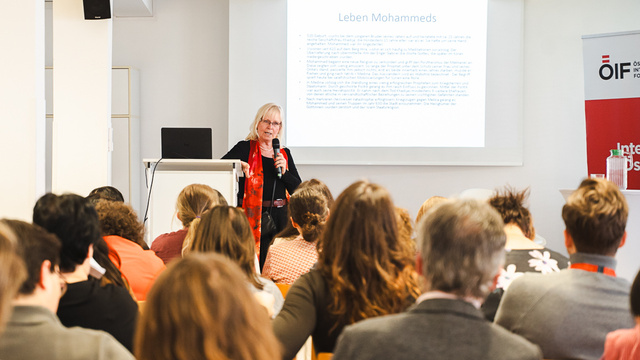 Eine Referentin steht an einem Pult und spricht vor einem Publikum in einem Seminarraum. Im Hintergrund ist eine Präsentationsfolie mit dem Titel „Leben Mohammeds“ zu sehen. Die Zuhörer sind größtenteils von hinten zu sehen und konzentrieren sich auf die Vortragende.