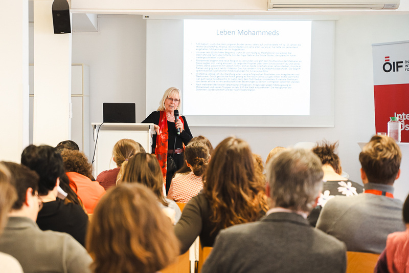 Eine Referentin steht an einem Pult und spricht vor einem Publikum in einem Seminarraum. Im Hintergrund ist eine Präsentationsfolie mit dem Titel „Leben Mohammeds“ zu sehen. Die Zuhörer sind größtenteils von hinten zu sehen und konzentrieren sich auf die Vortragende.