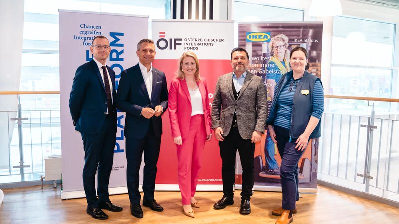 Fünf Personen stehen nebeneinander vor Werbebannern für Integrationsprojekte und Arbeitsmöglichkeiten.
