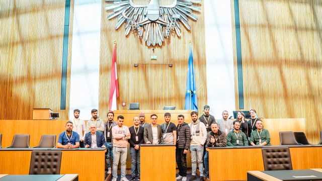 Eine Gruppe von Menschen steht im Sitzungssaal mit einem Adler-Wappen im Hintergrund.