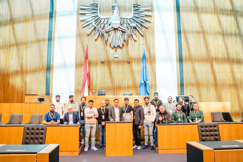 Eine Gruppe von Menschen steht im Sitzungssaal mit einem Adler-Wappen im Hintergrund.